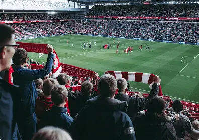 A view of a football pitch from the stands. Fans holding up their team scarves are cheering on the game about to start.